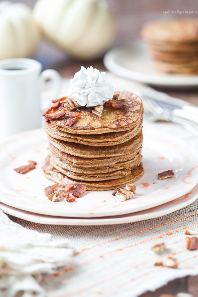 A stack of delicious spiced pumpkin pancakes topped with cooked bacon, chopped pecans and whipped cream.