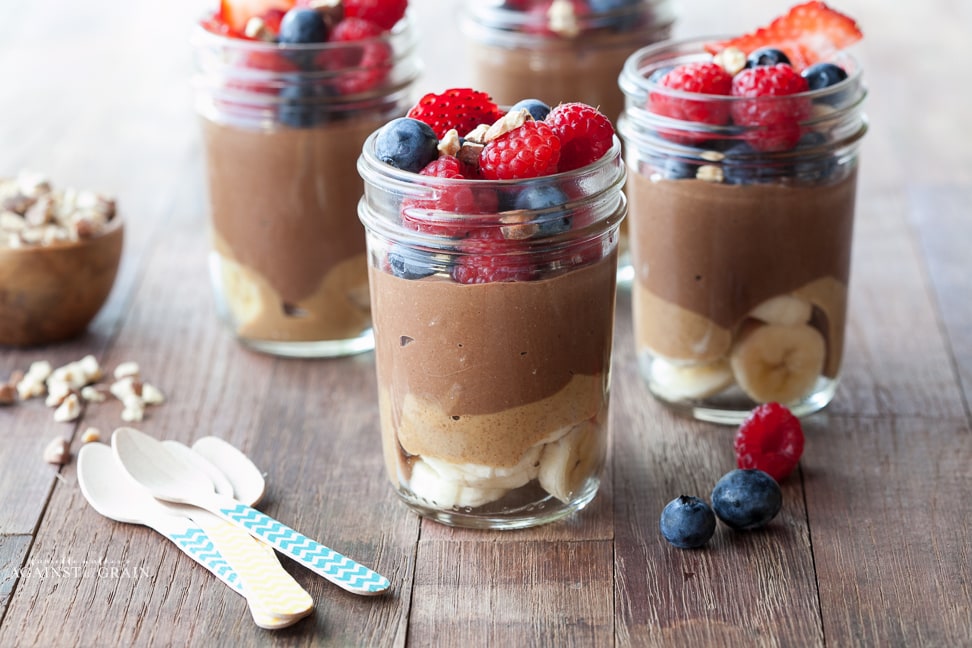 4 mason jars filled with chia/chocolate pudding with almond butter and bananas on bottom and fresh berries on top.