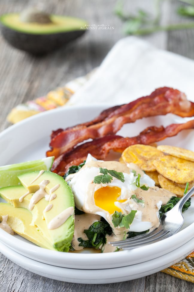 Whole30 friendly Mexican eggs benedict served with fresh avocados, plantains and bacon on a white plate placed on a wooden table. 