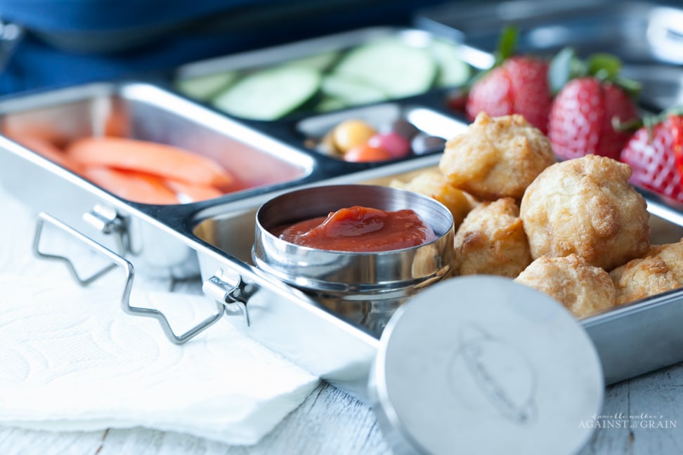 A lunch box filled with chicken nuggets, veggies, fruit and a container of homemade paleo ketchup. The ketchup is sweetened with dates and kid approved!