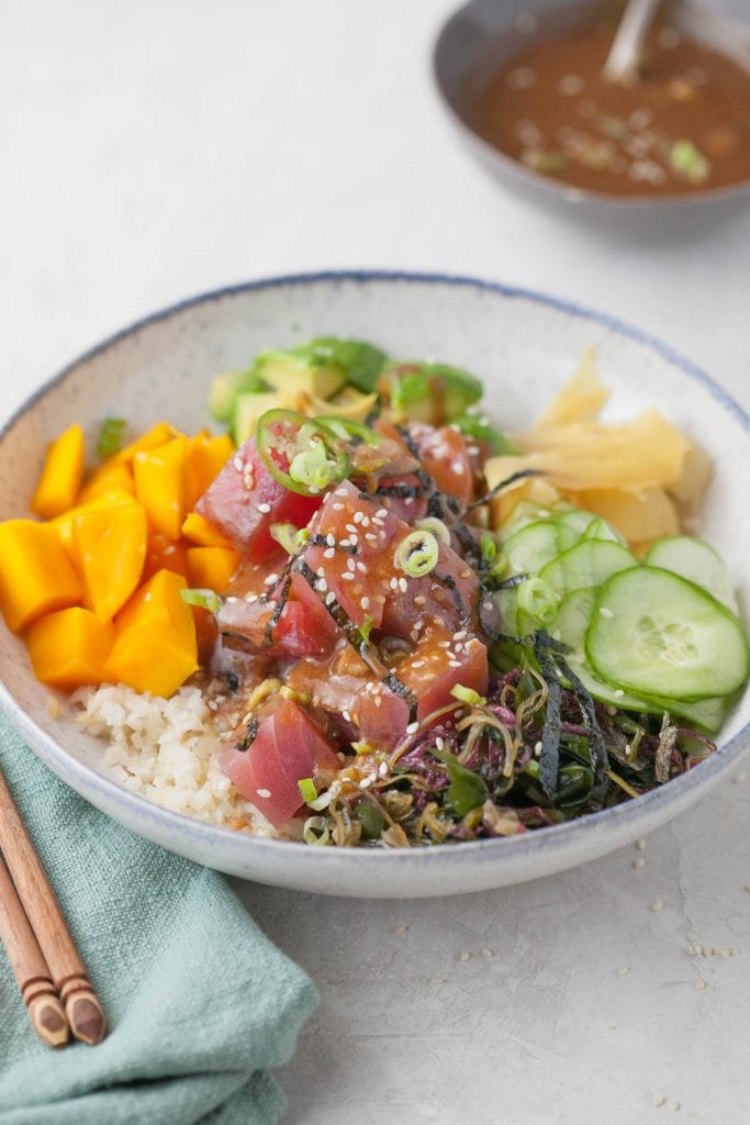 poke bowl with raw tuna, cauliflower rice, avocado, ginger, mango and thin slices of cucumber with homemade sauce drizzled on top