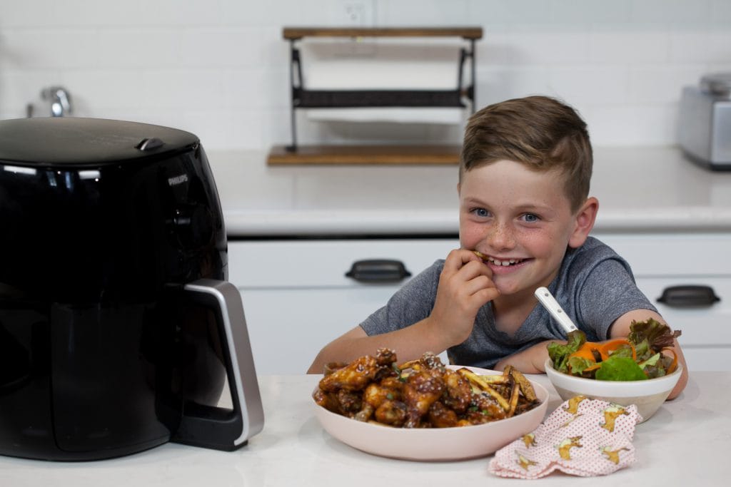 young boy smiles at camera waiting to dive into a bowl of Sticky Sesame Chicken Wings with french fries while he stands in home kitchen next to an air fryer