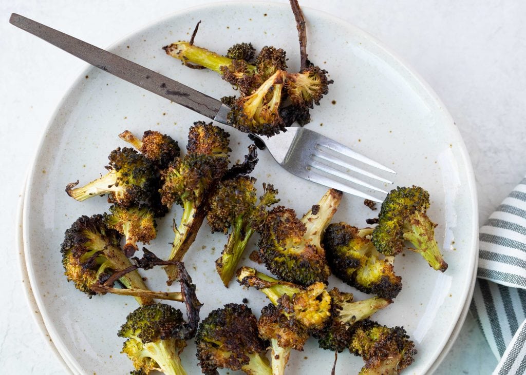 well seasoned roasted broccoli on a white plate with a silver fork on white counter. A blue and white striped hand towel lays next to plate.