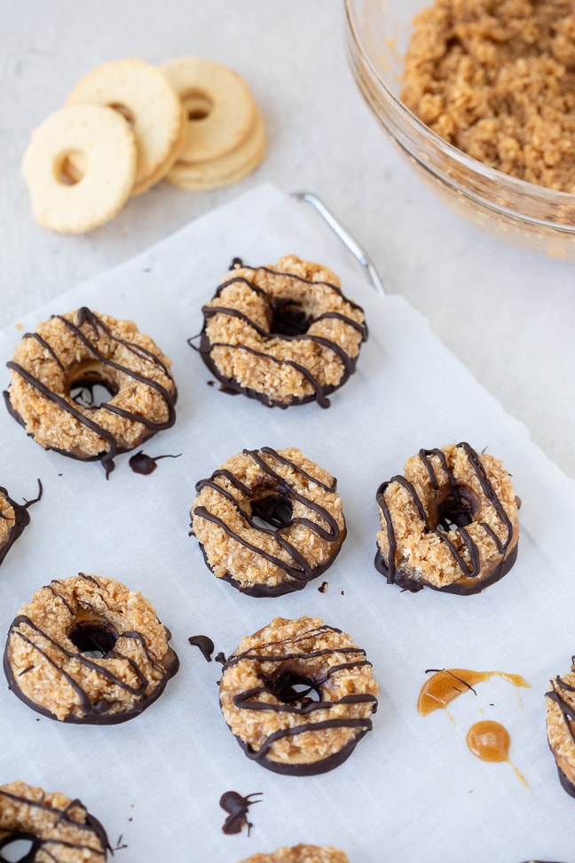 On a white surface; view of homemade samoa cookies with chocolate drizzle on a cooling rack with parchment paper. In the image, shortbread cookies and coconut mixture in clear bowl is visible. 