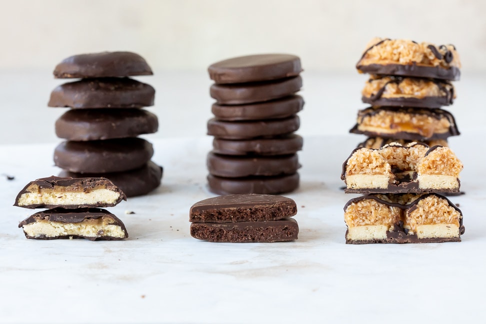 A variety of girl scout cookie renditions (tagalong, thin mint and samoas) stacked on a white surface. Each stack has a single cookie cut in half in front of it to display inside. 