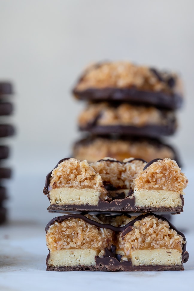 up close view of samoa cookie cut in half with a blurred view of a cookie stack in the background; all on a white surface. 