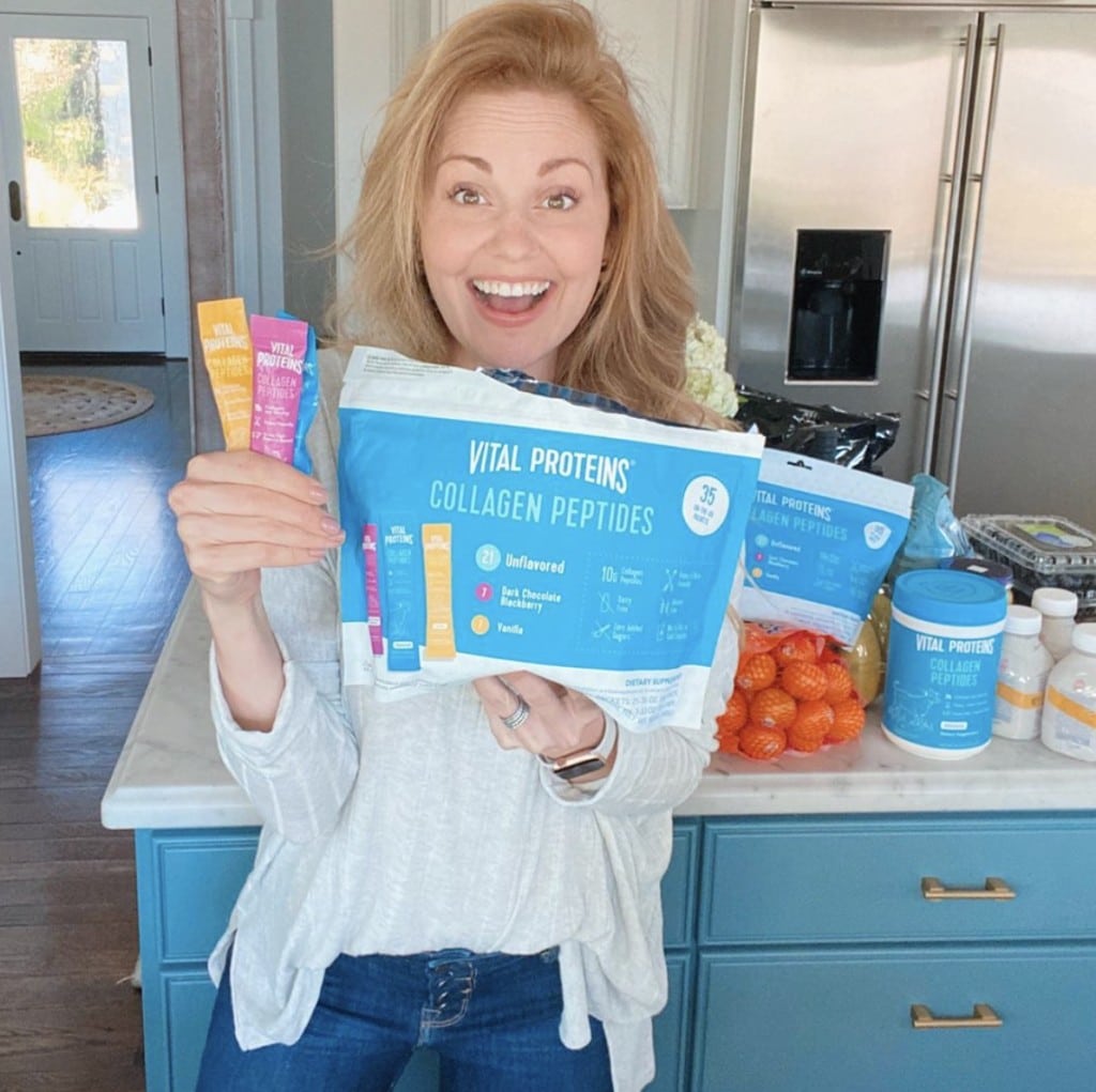 Smiling woman looking into camera holding packages of Vital Proteins in home kitchen