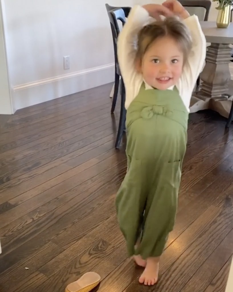 female child wearing an olive green jumpsuit practices ballet while barefoot in her home