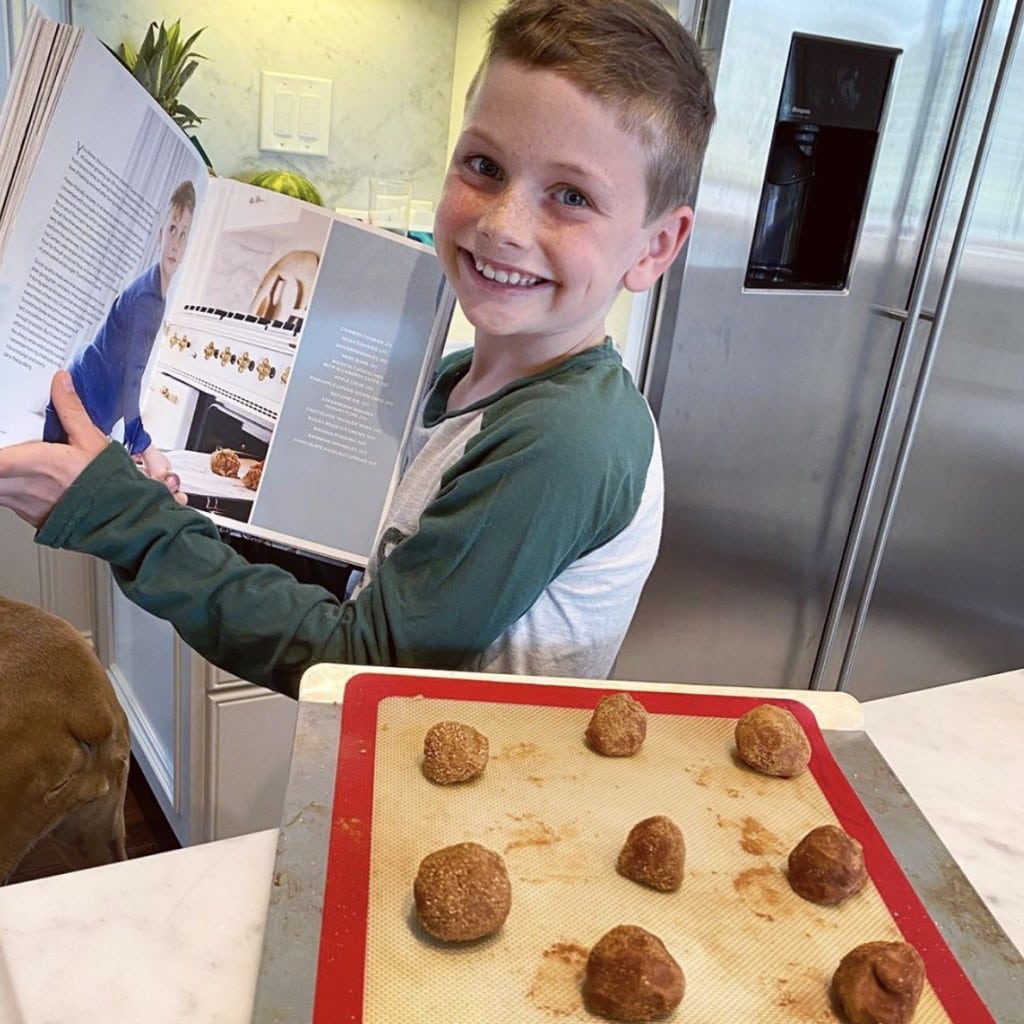 Beaming male child proudly holds up cookbook while displaying his made from scratch cookies prior to baking while standing in home kitchen