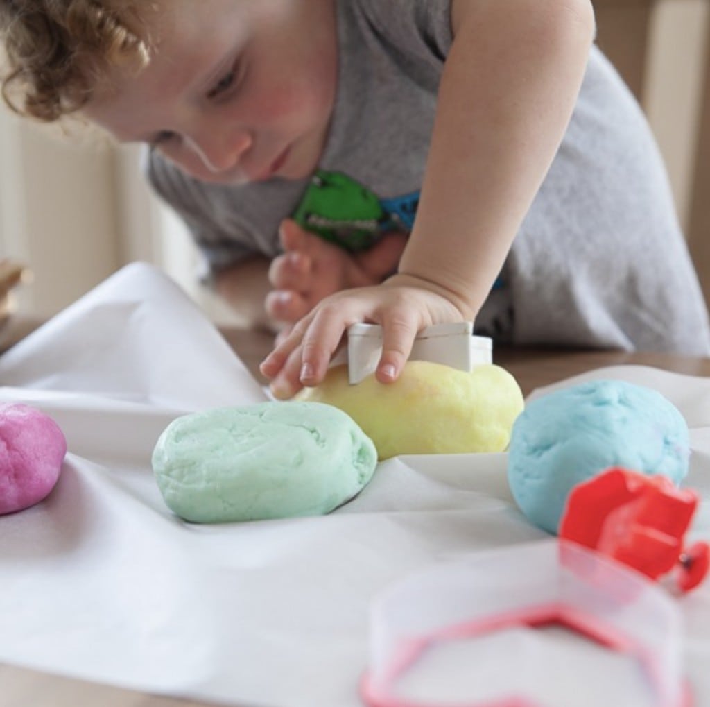 small male child with curly hair uses a cookie cutter to cut into a variety of pastel colored play dough