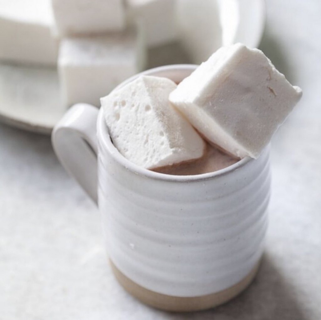 white cup of hot chocolate topped with marshmallows on a white counter with a platter of marshmallows in the background