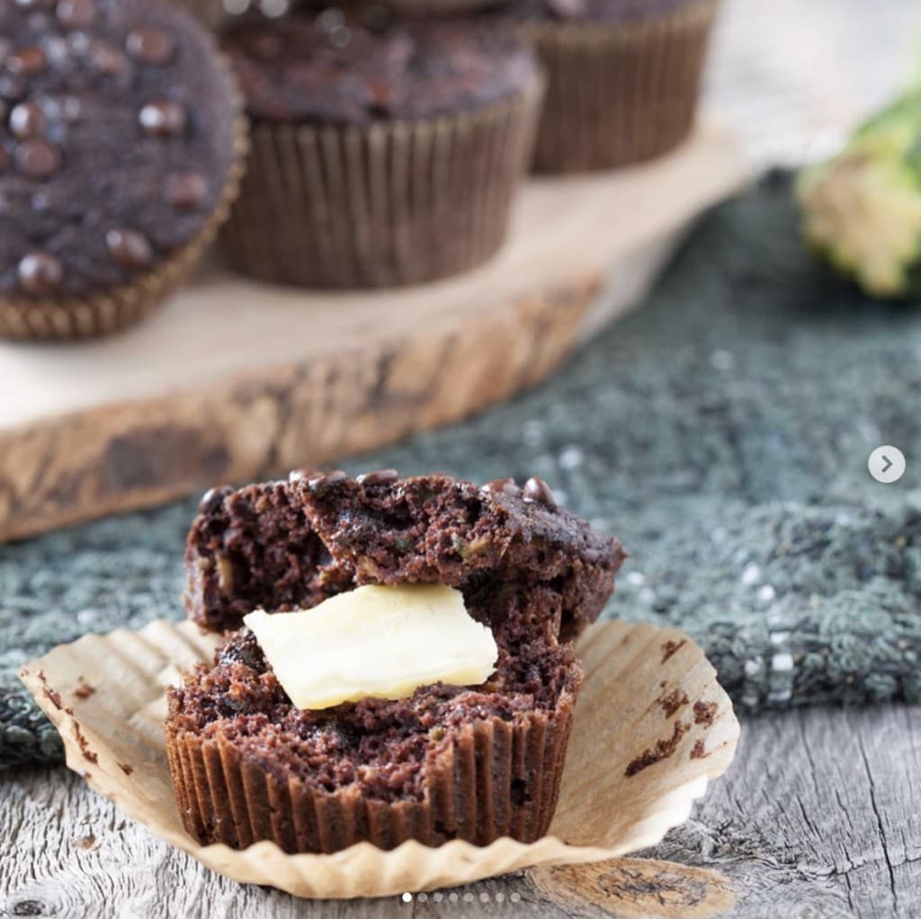 chocolate muffin in parchment cupcake wrapper cut in half with butter in the middle on a wooden surface with a display of chocolate muffins in the background on a wooden platter 