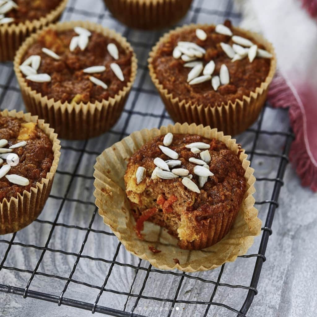 brown muffins topped with sunflower seeds in parchment paper cupcake wrappers cooling on a wire rack on a light gray wooden surface 