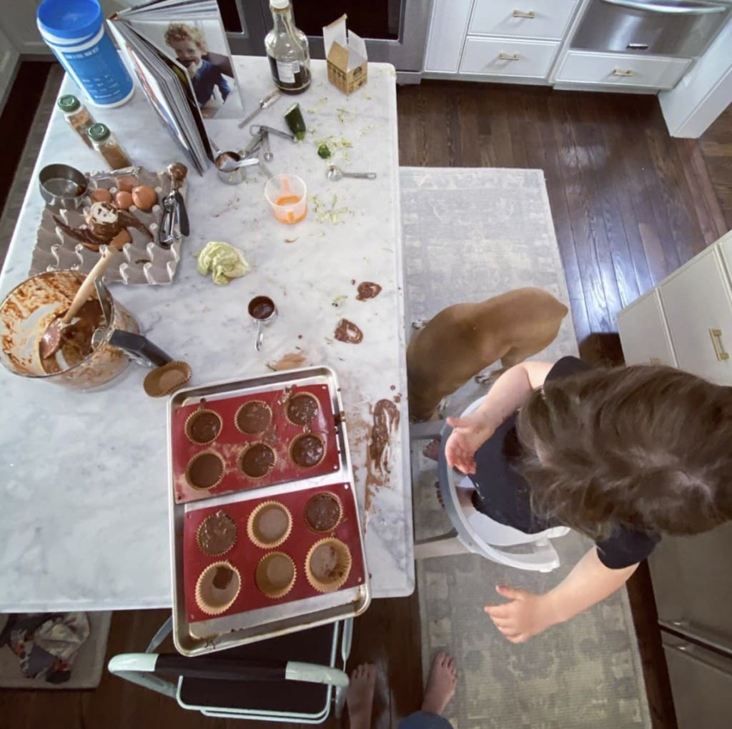 young girl stands on chair at kitchen counter preparing muffins. The counter is a general disarray with various baking ingredients and cookbook