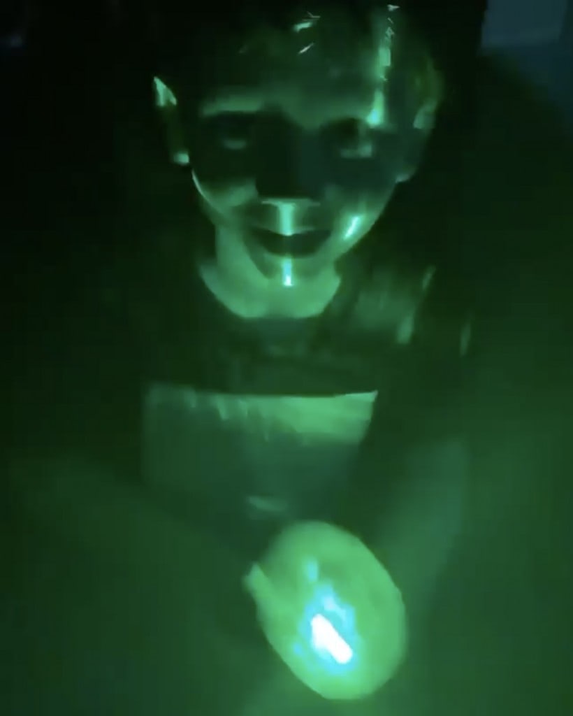 young boy smiles at camera while he plays with a green flashlight in the dark