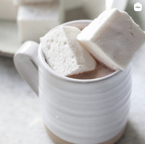 hot chocolate topped with two marshmallows in a white cup with a platter of marshmallows in the background on a white marble counter