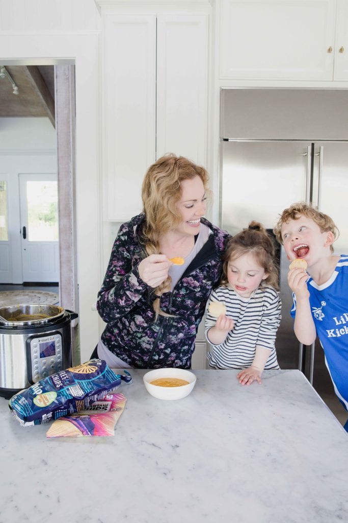 In a home kitchen, by an Instant Pot and open bags of chips, woman smiles at children, a young boy and young girl. Young boy and woman and playfully smiling at each other while holding chips and young girl looks at her chip. 