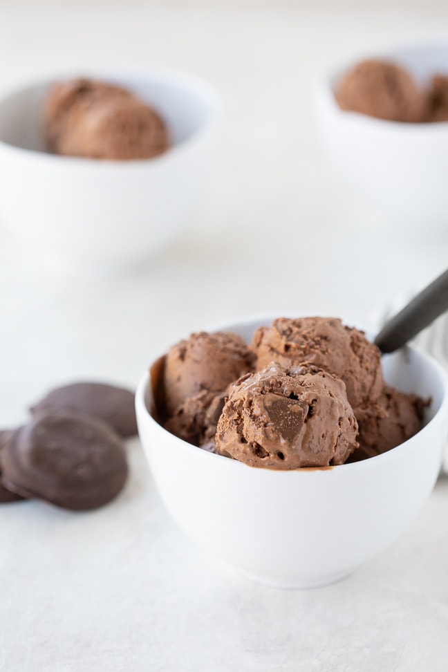 white bowl of double chocolate thin mint ice cream on a white surface. Thin mint cookies and additional ice cream filled bowls in background