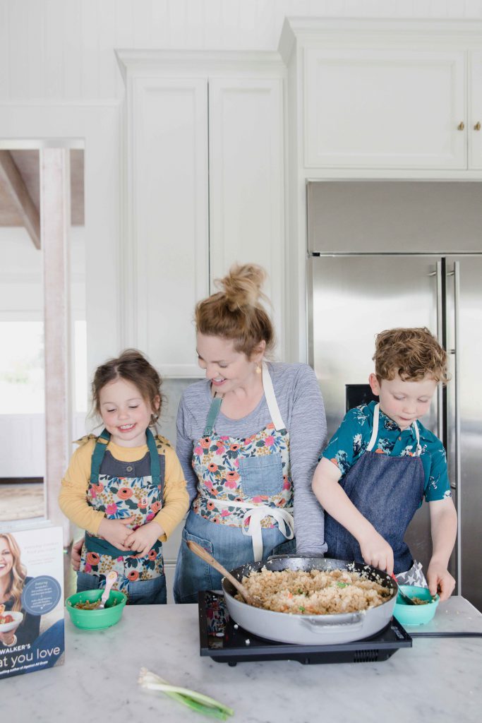 woman and children stand over skillet placed on counter top, smiling and cooking. Cookbook Eat What You Love is displayed on counter