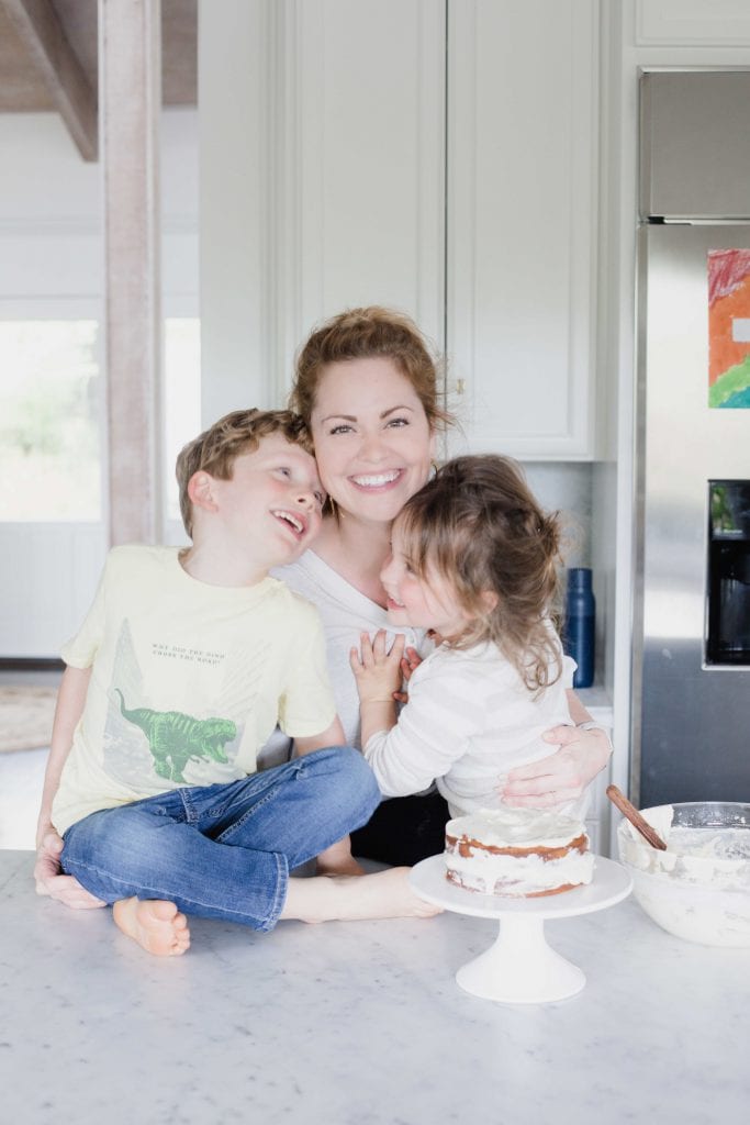 woman smiles at camera while embracing two happy children while standing in front of a counter with a partially decorated cake