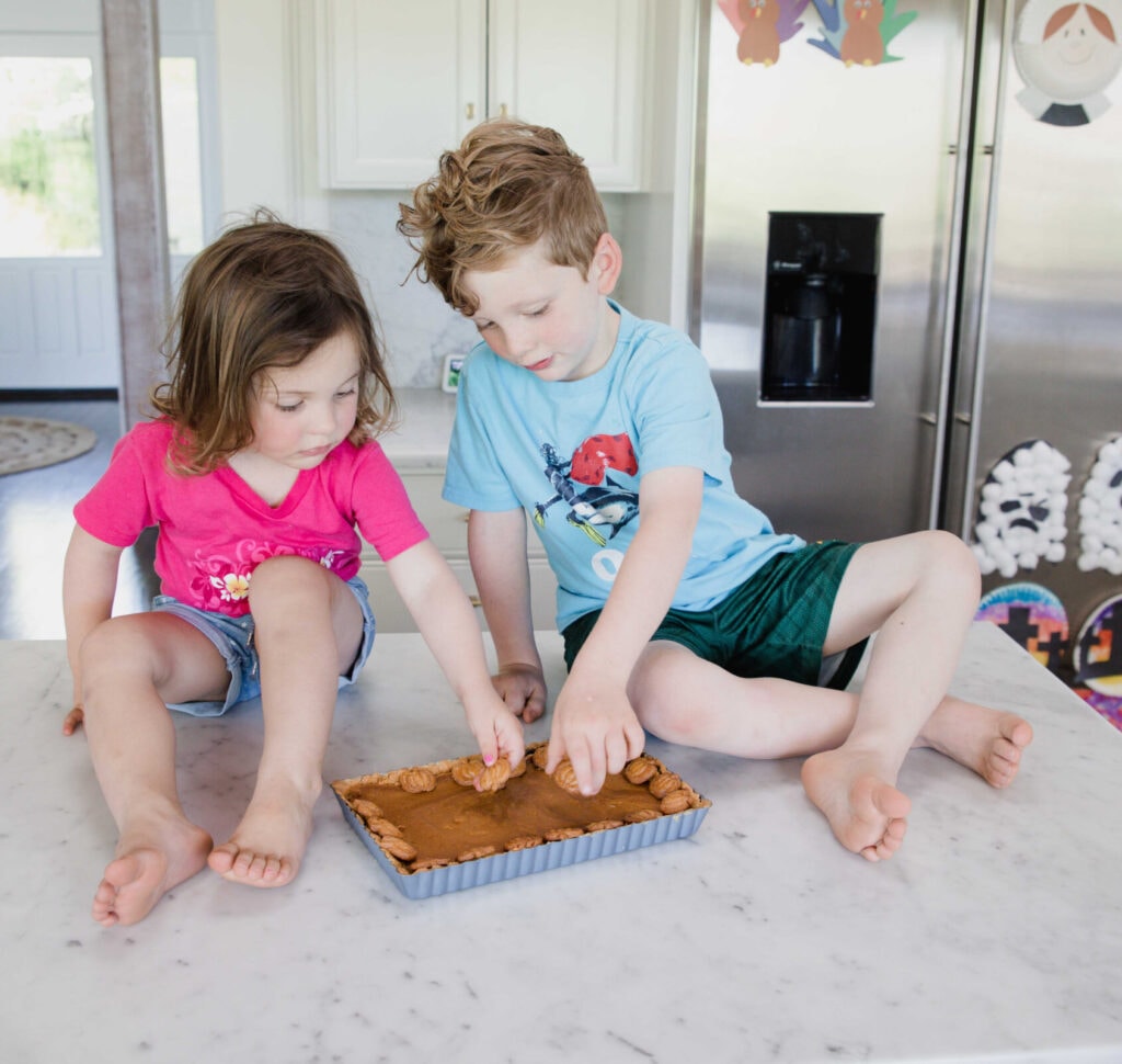 two children sit on a white marble counter decorating a pumpkin pie