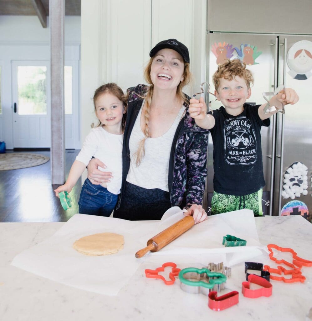 Mother and children smile at camera while standing at counter with freshly made sugar cookie dough and Christmas cookie cutters