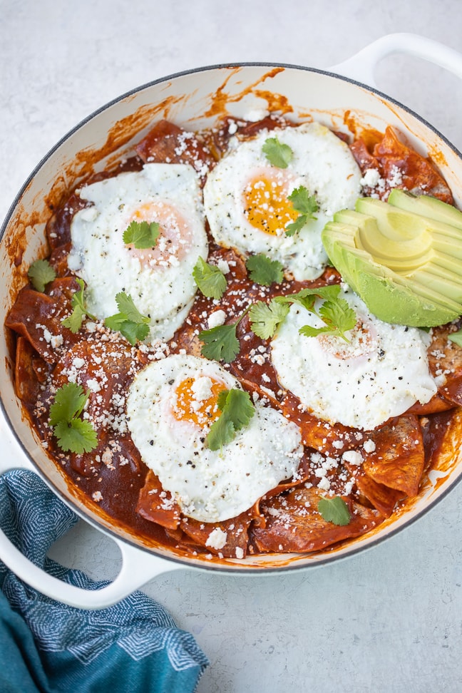 a pot of grain and gluten-free chilaquiles; chips smothered in red sauce with four eggs over easy on top with a side of avocado with cilantro as garnish