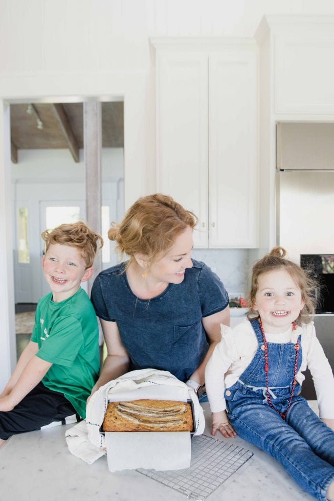 Young boy and girl sit on kitchen counter smiling at camera while woman displays freshly baked banana bread