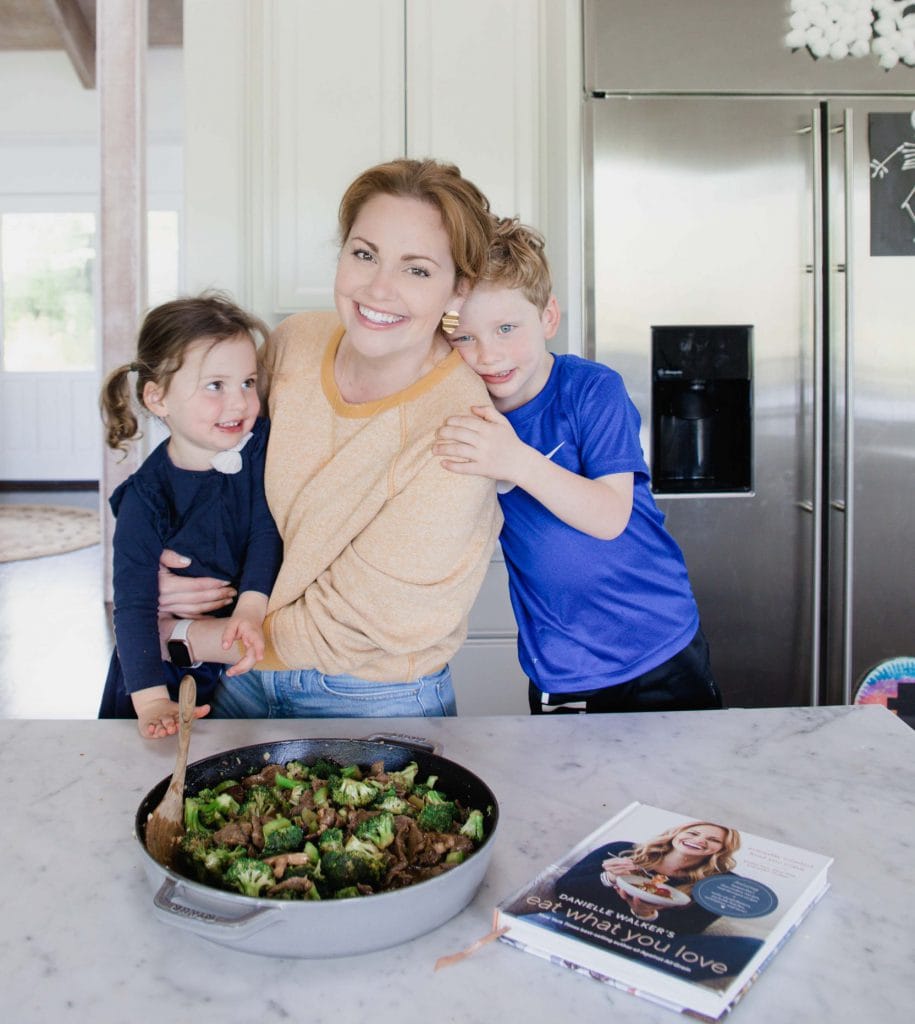 Woman stands in home kitchen embracing her daughter while she is embraced by her son. They are all smiling. On the counter rests a pot of beef and broccoli stir fry and her cookbook lays close by. 