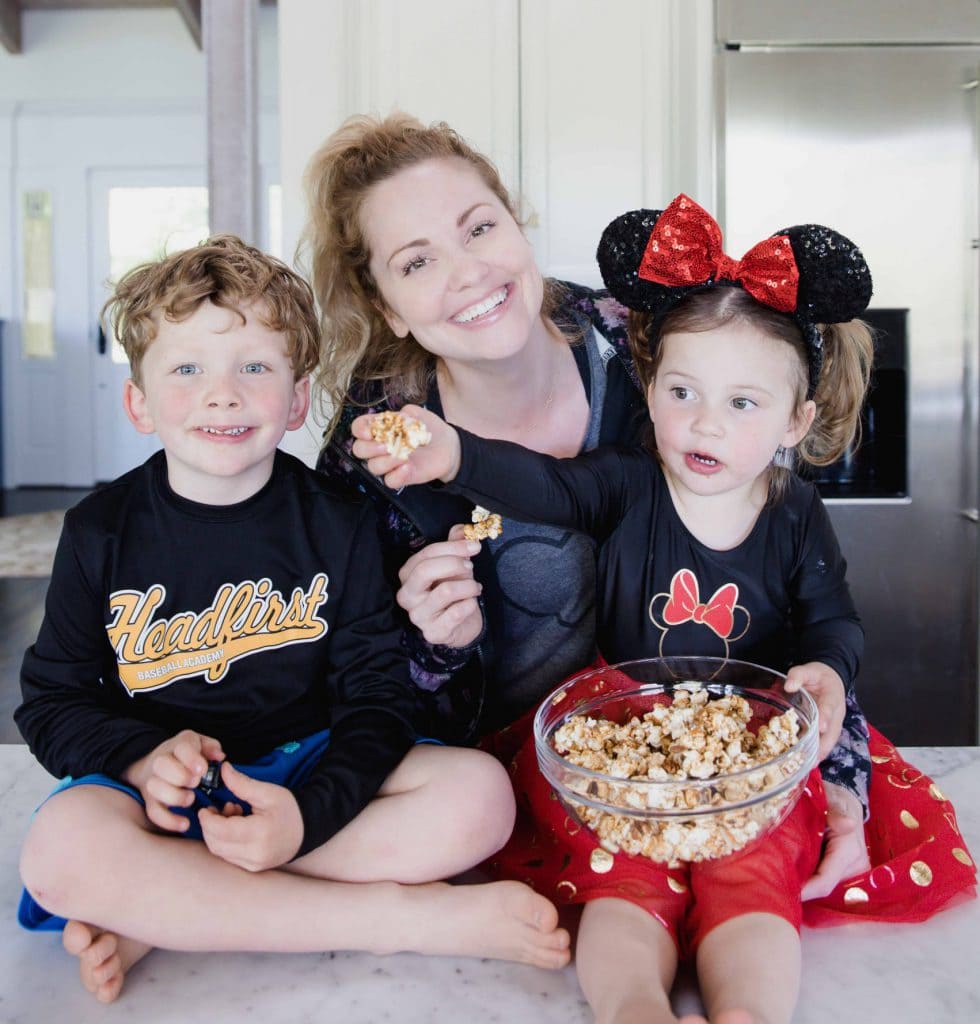 woman smiles at camera while standing behind children in home kitchen. The young girl holds a bowl of caramel corn. 