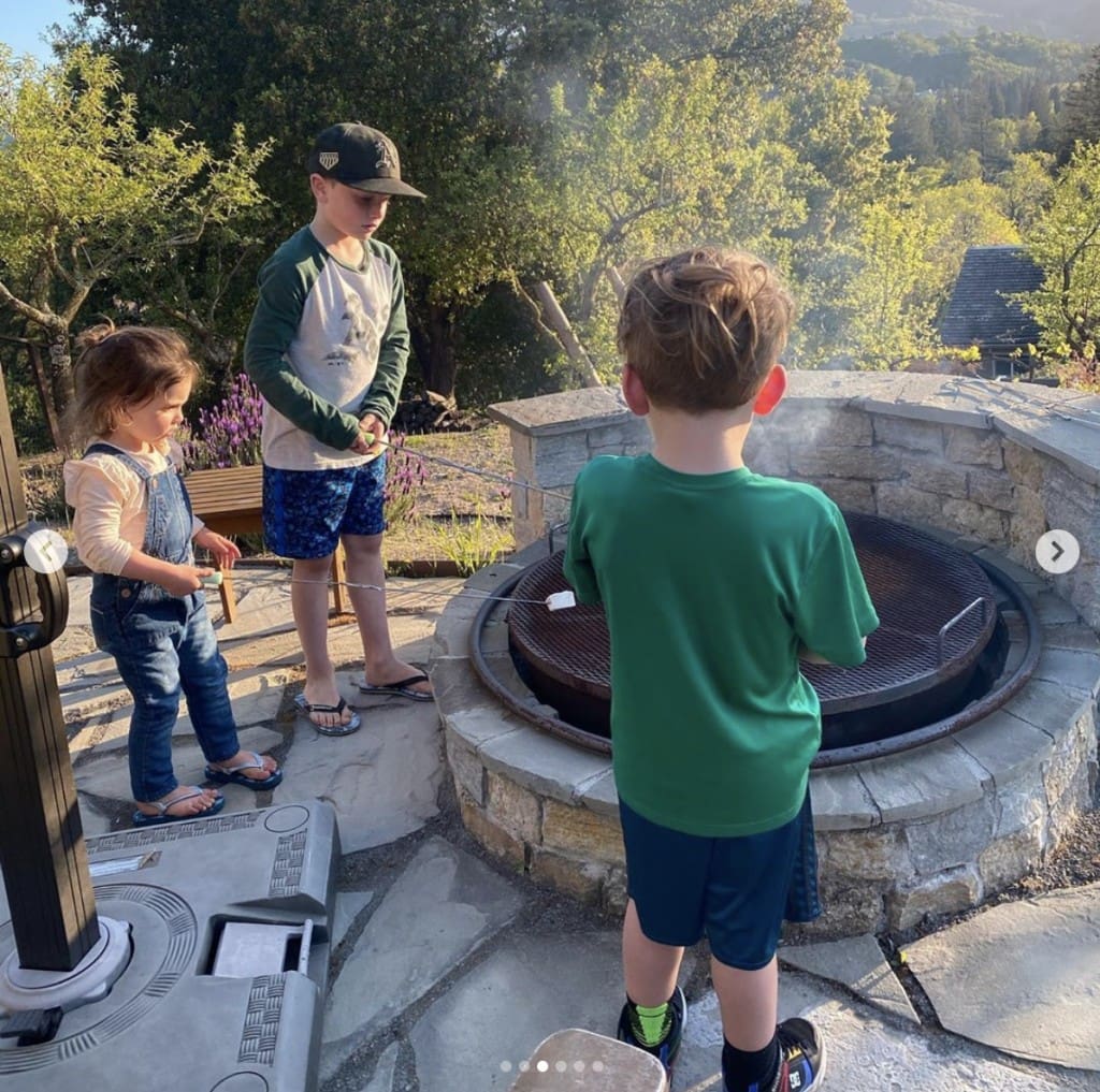 one young girl and two young boys gather around a stone fire pit to roast marshmallows