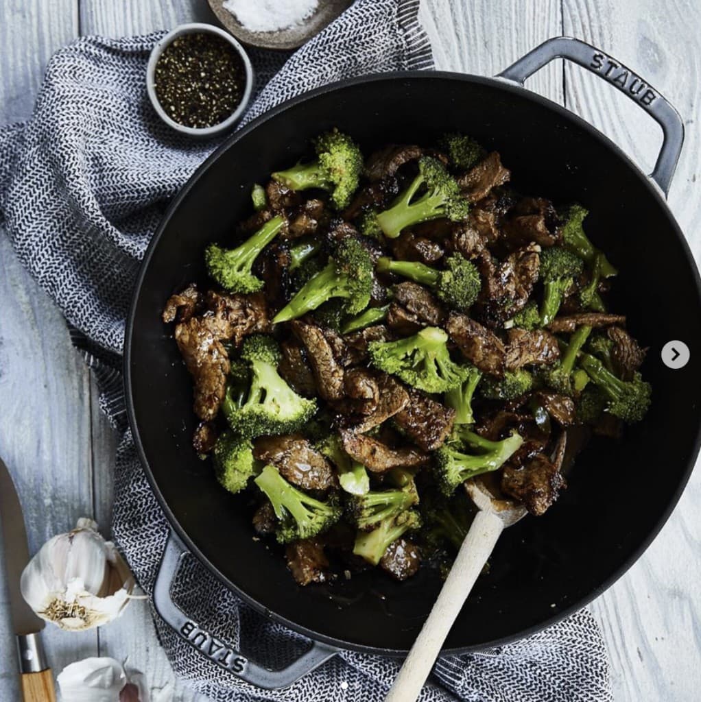 cooked beef and broccoli in a black Staub pot on a gray wooden table. Salt, pepper and garlic cloves are on the side. 