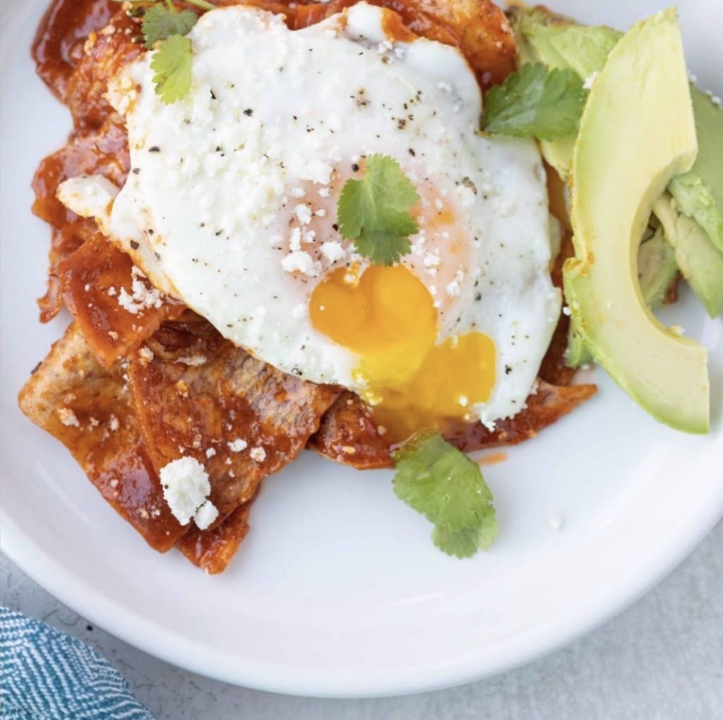 fried egg with a runny yolk lays on top of chips smothered in sauce with a side of fresh avocado on white plate