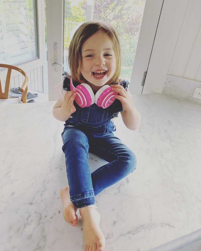 young girl sits on counter smiling gleefully at camera while holding pink headphones around her neck