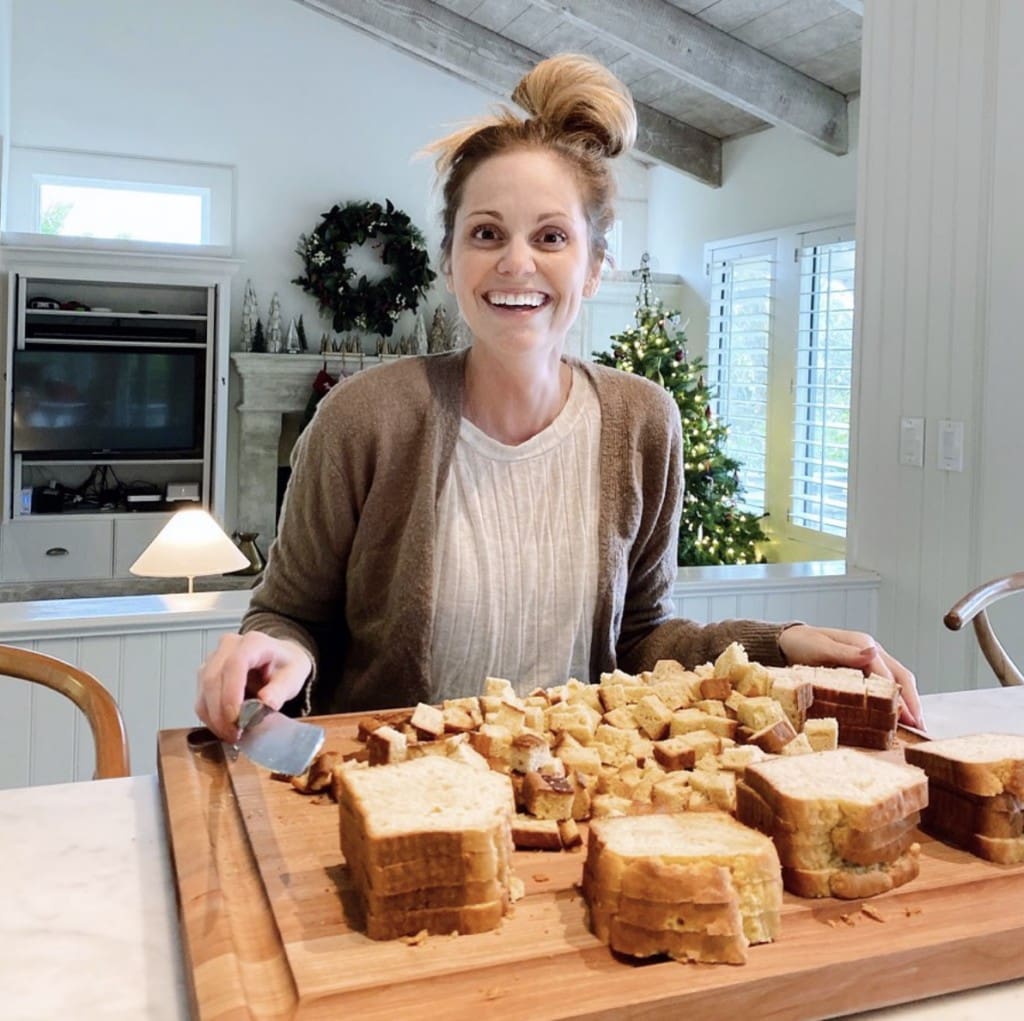 woman sits at counter smiling at camera; she is dicing up slices of bread to prepare stuffing