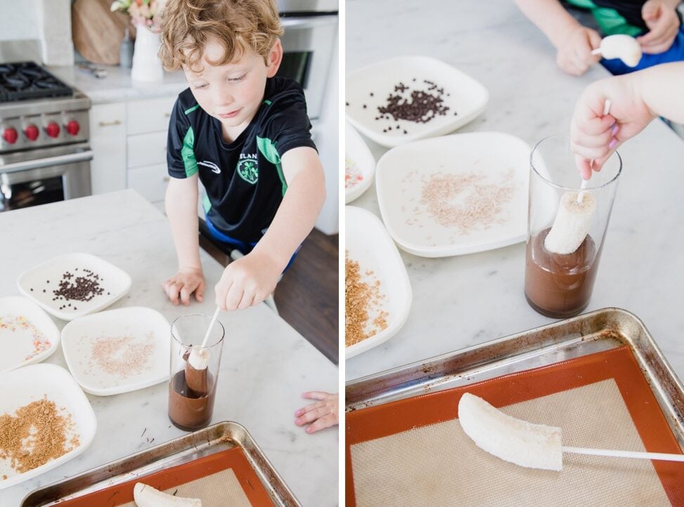 young boy carefully dips frozen bananas into melted chocolate in his home kitchen. A variety of toppings are on separate plates on the counter. 