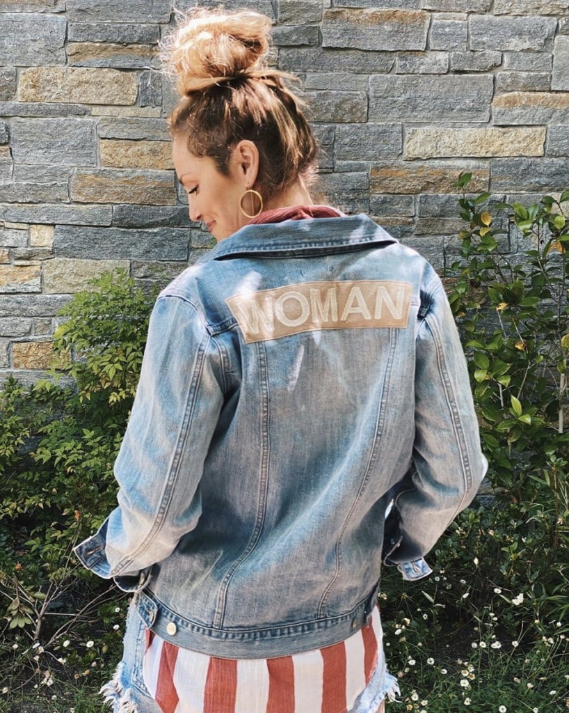 woman stands with back to camera modeling light blue denim jacket that has the word "woman" in all caps embroidered