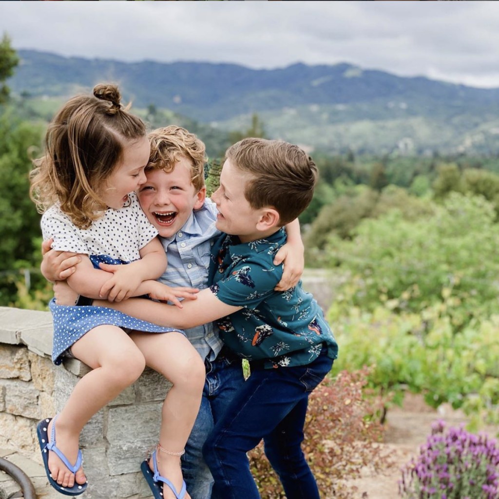 Three children embrace each other while they laugh lovingly against a backdrop of picturesque rolling green hills