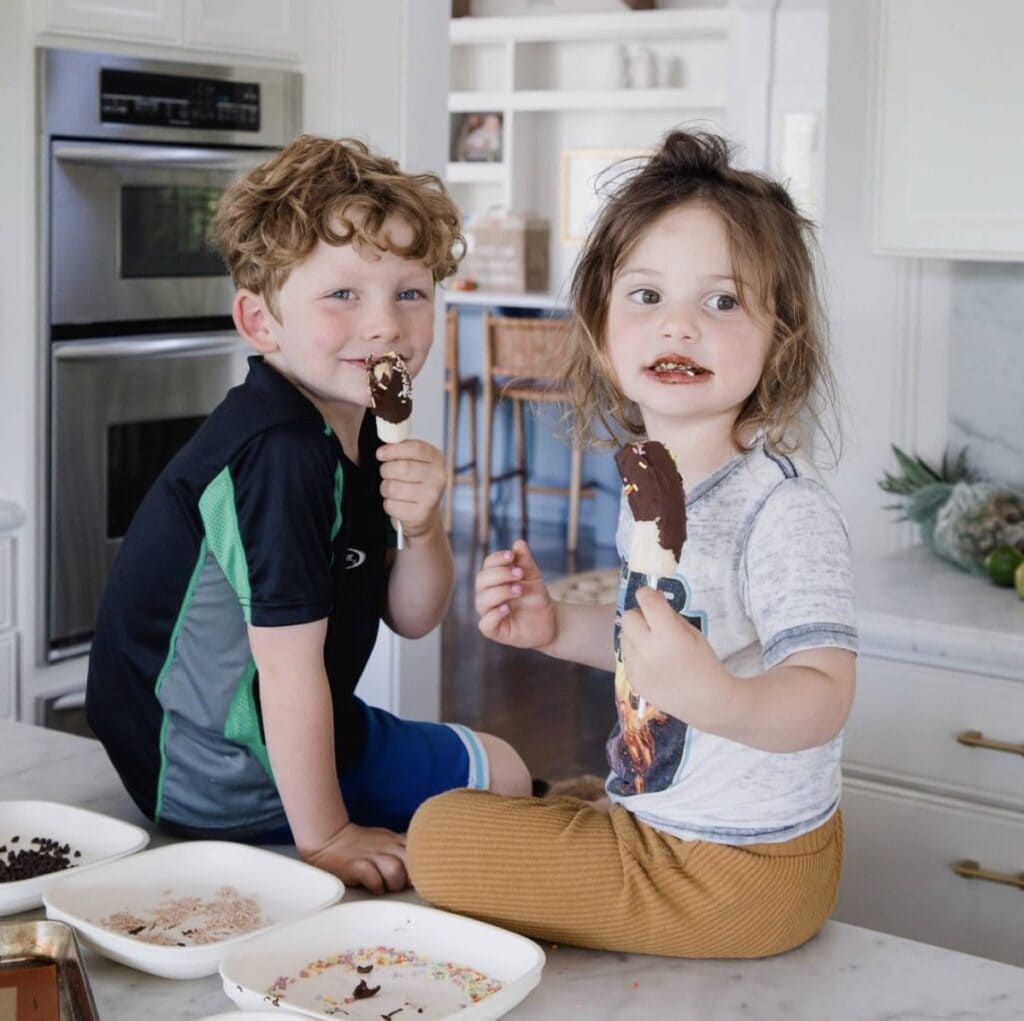 young male and female child sit on kitchen counter enjoying frozen chocolate dipped bananas