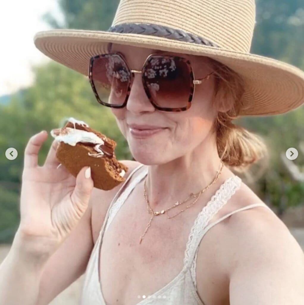 Woman wears sunhat and sunglasses and smiles at the camera while holding a s'more