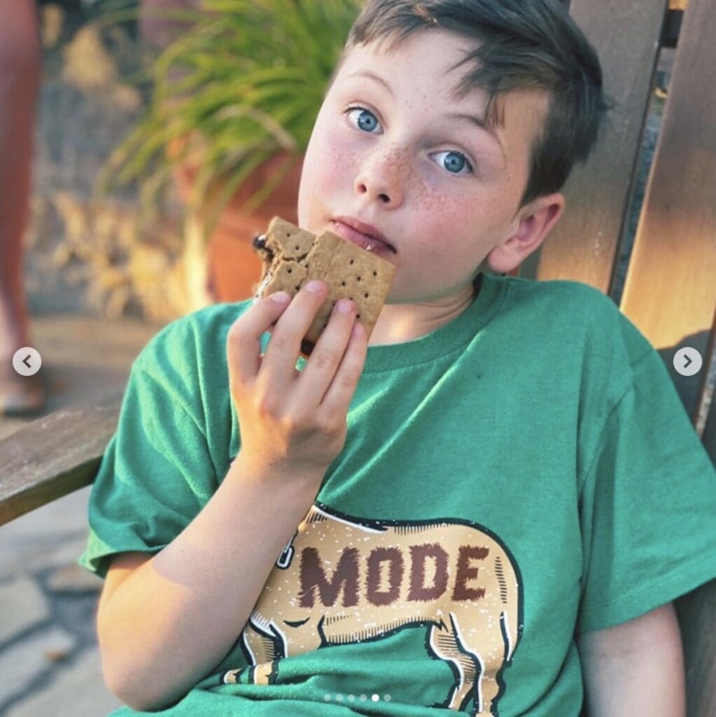 Boy wearing a green t-shirt stares seriously into camera as he gets ready to bite into his s'more 