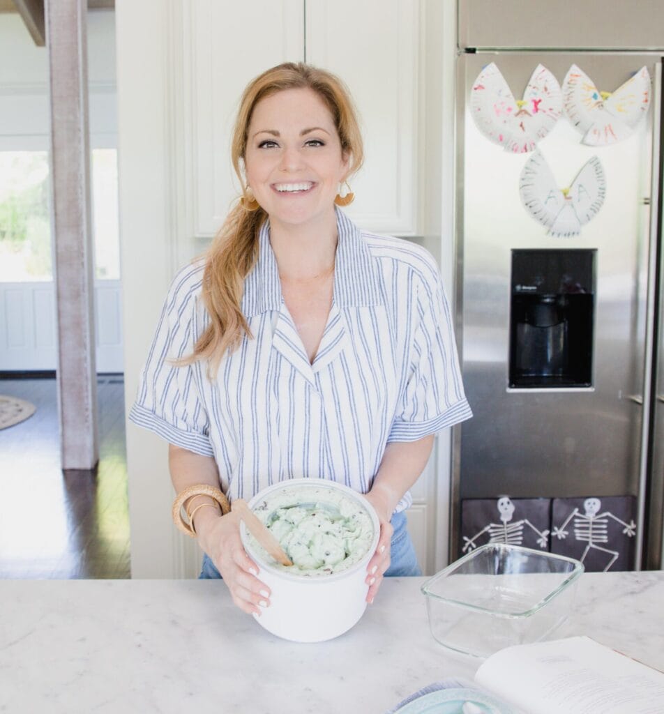 Woman stands in home kitchen smiling at camera while holding a tub of freshly made mint chocolate chip ice cream