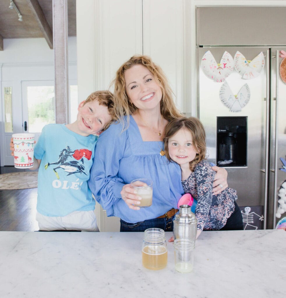 Woman stands in home kitchen smiling at camera while holding a drink in one hand & embracing her young daughter with the other. Her young son holds a kid friendly drink in his hand and leans against her as he smiles at the camera as well. 