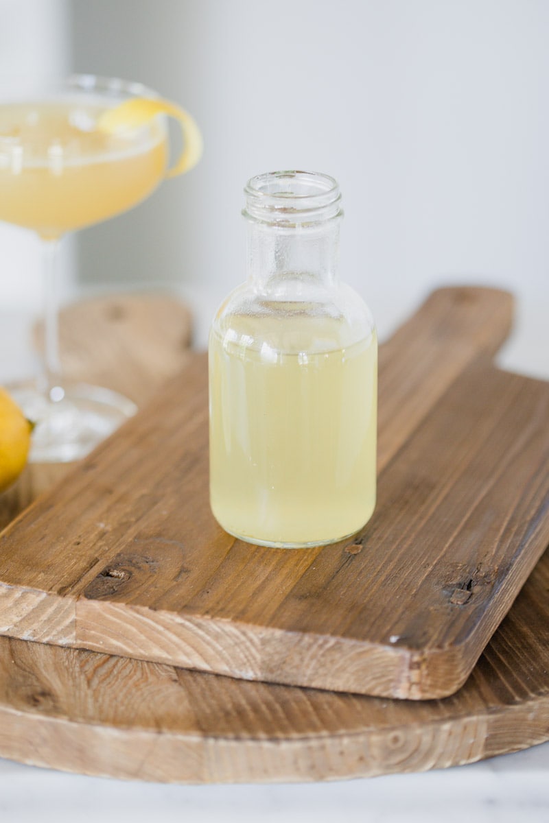 honey simple syrup in a small glass bottle resting on wooden cutting boards with a yellow colored cocktail in the background