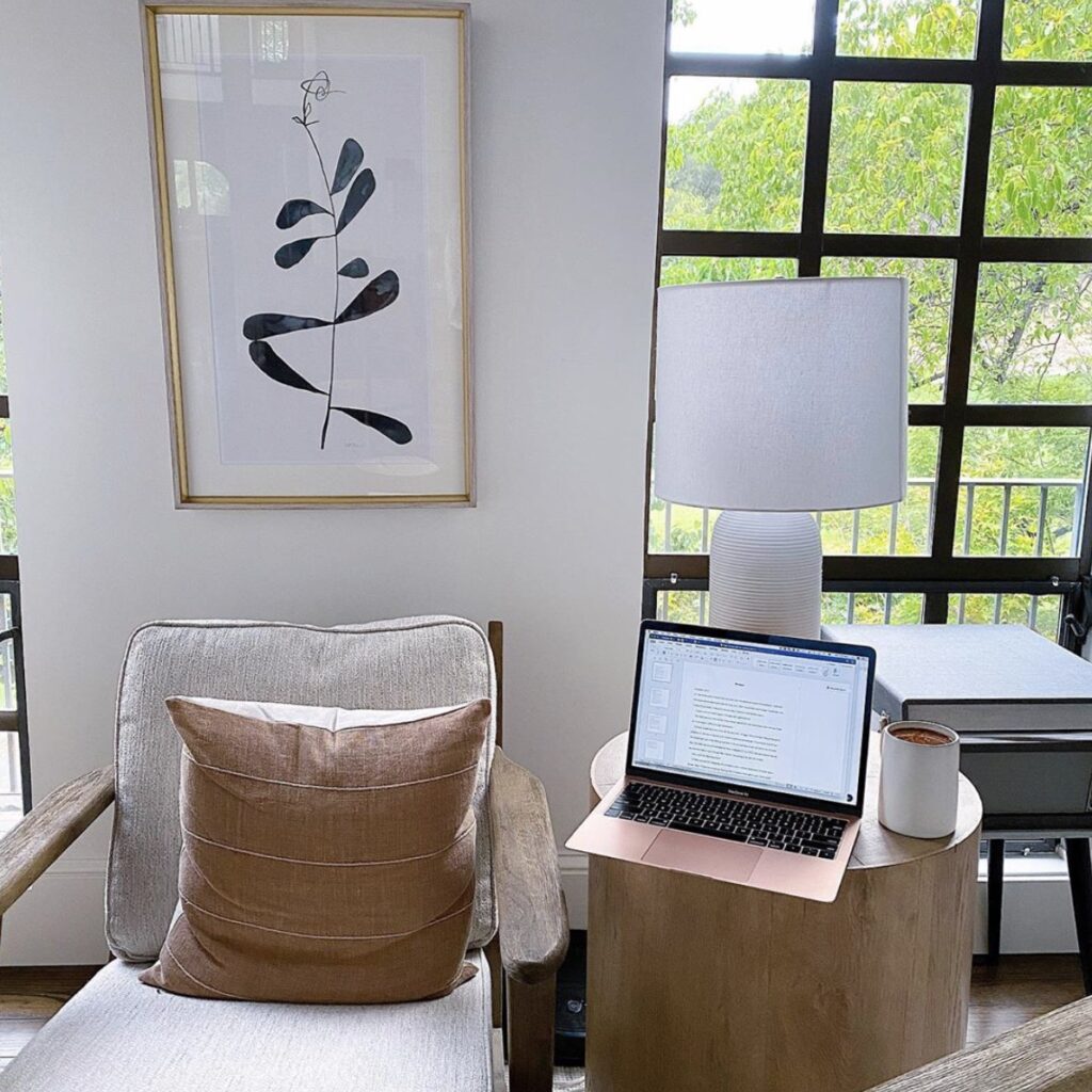 tastefully adorned living space by a window. Laptop and cup of freshly brewed coffee sit on a round wooden lamp stand