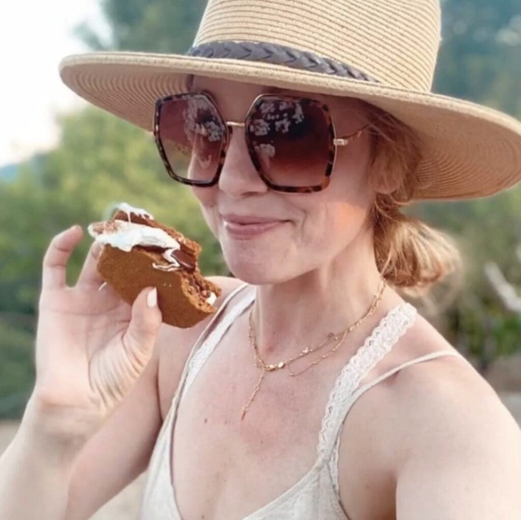 woman holds a s'more up to the camera and smiles mischievously. She wears oversized sunglasses and a sunhat. 