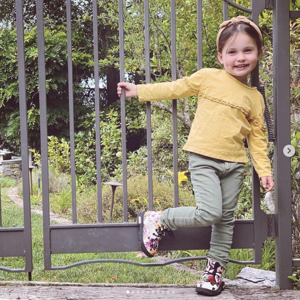 Young girl poses next to fence wearing floral combat boots, olive green jeans and a mustard yellow shirt with matching headband. She smiles directly at camera. 