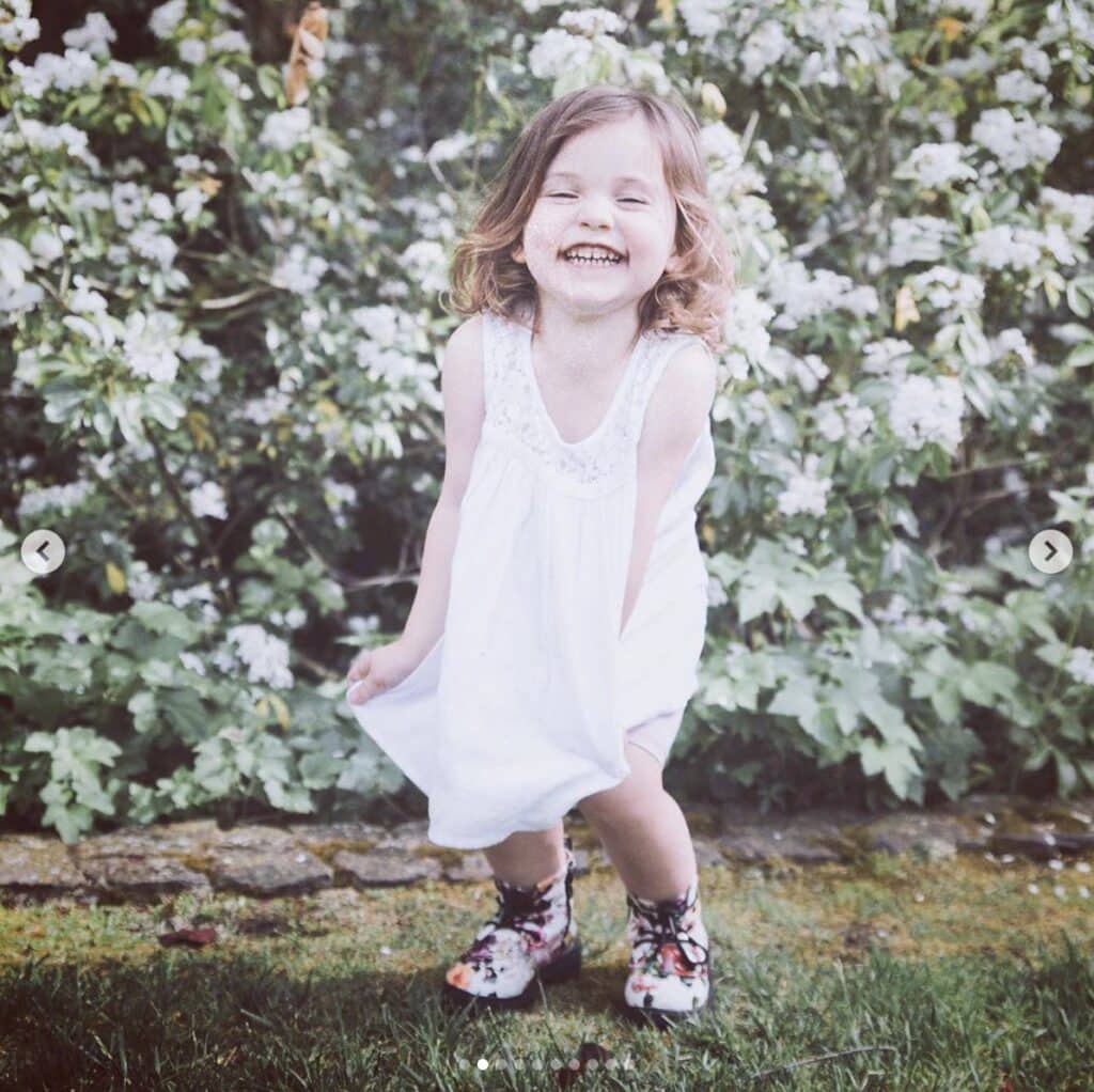 young girl laughs in front of floral bush outdoors. She's wearing floral combat boots and a white dress