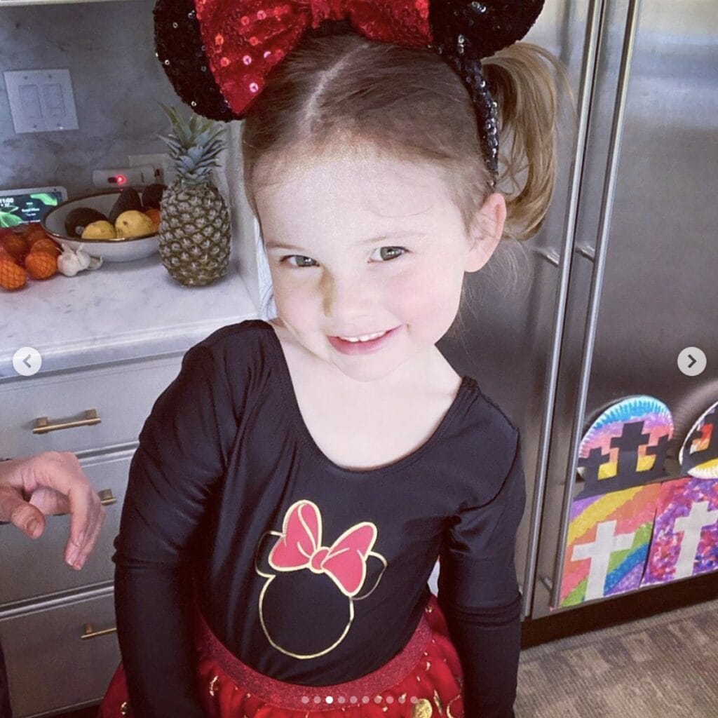 Young girl smiles at camera while standing in home kitchen. She wears a black and red Minnie Mouse tutu dress, complete with sequined ear headband