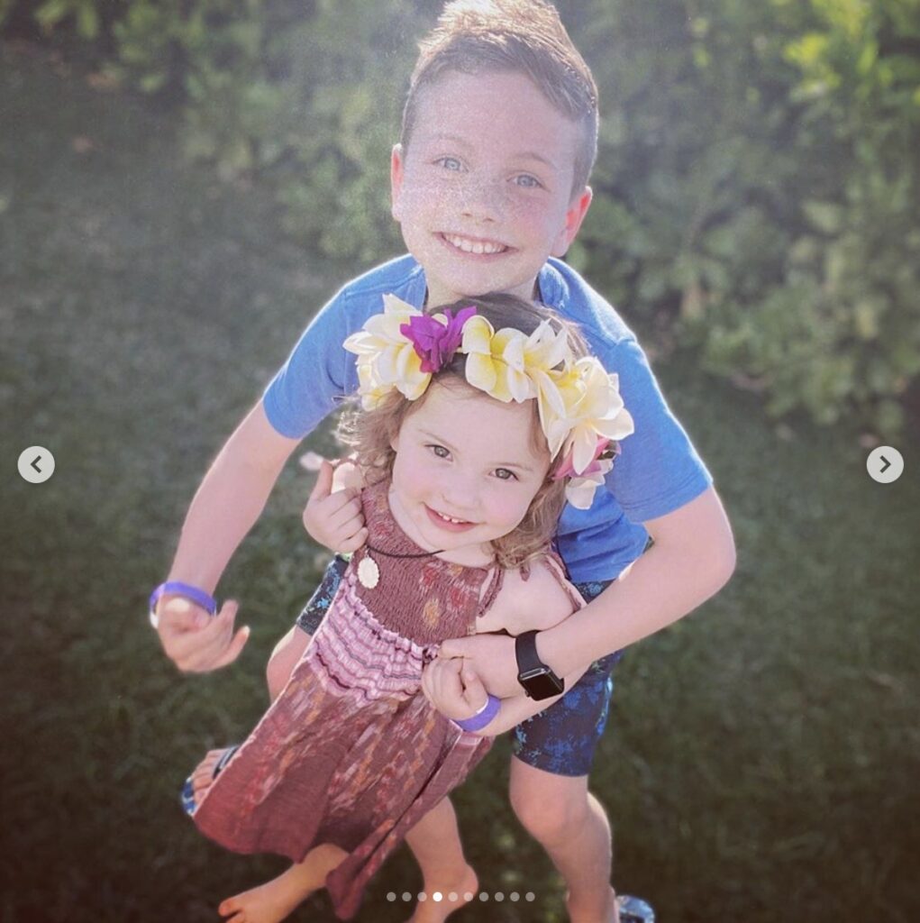 young girl wearing a flower crown is embraced by older brother. They both smile directly at the camera. 